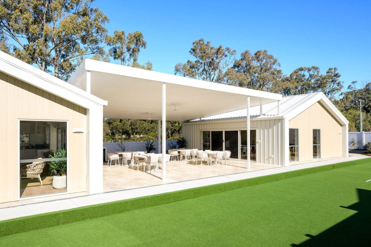 A modern building with a spacious covered patio featuring tables and chairs, large windows, light-coloured exterior, and an artificial grass lawn—perfect for enjoying the Hunter Valley over 50s neighbourhood lifestyle amid tall trees and blue skies.