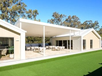 A modern building with a spacious covered patio featuring tables and chairs, large windows, light-coloured exterior, and an artificial grass lawn—perfect for enjoying the Hunter Valley over 50s neighbourhood lifestyle amid tall trees and blue skies.