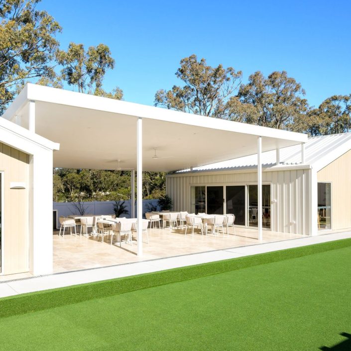 A modern building with a spacious covered patio featuring tables and chairs, large windows, light-coloured exterior, and an artificial grass lawn—perfect for enjoying the Hunter Valley over 50s neighbourhood lifestyle amid tall trees and blue skies.