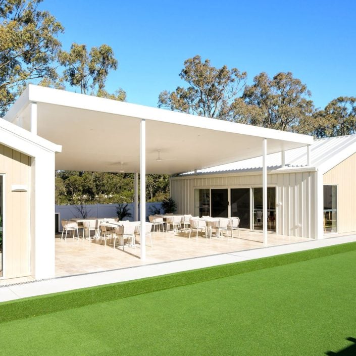 A modern building with a spacious covered patio featuring tables and chairs, large windows, light-coloured exterior, and an artificial grass lawn—perfect for enjoying the Hunter Valley over 50s neighbourhood lifestyle amid tall trees and blue skies.