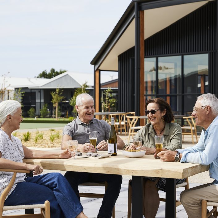 Four older adults sit at an outdoor table in a retirement living community, smiling and talking whilst enjoying drinks and snacks. Modern buildings and greenery are visible in the background on a sunny day.