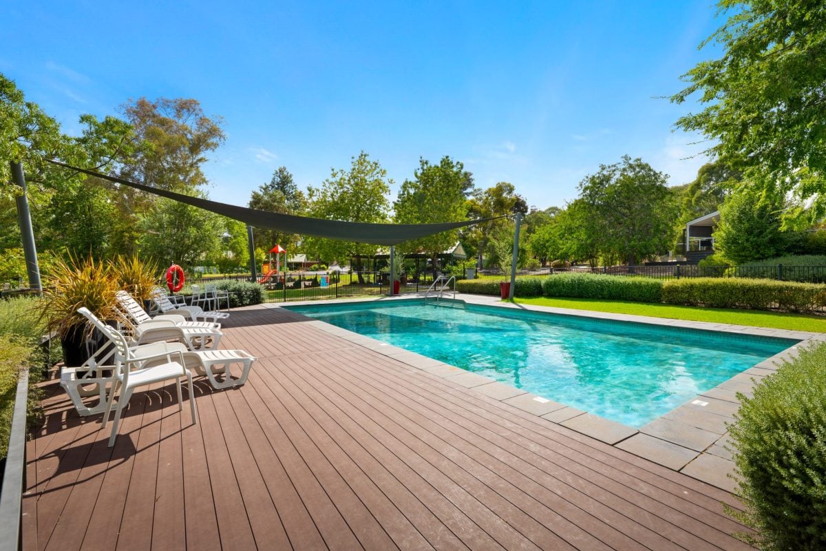Outdoor swimming pool with clear blue water, surrounded by a wooden decking and white loungers—perfect for Albury retirement over 50s. Trees and greenery frame the scene under a sunny sky, with a large sunshade stretched over one end.