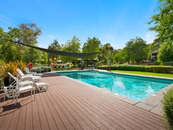Outdoor swimming pool with clear blue water, surrounded by a wooden decking and white loungers—perfect for Albury retirement over 50s. Trees and greenery frame the scene under a sunny sky, with a large sunshade stretched over one end.