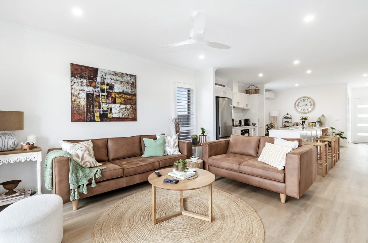 Modern living room with two brown sofas, a round wooden coffee table on a woven rug, kitchen in the background, and abstract artwork on the wall; ideal for land lease living or retirement in bright, neutral tones and cosy decor throughout.