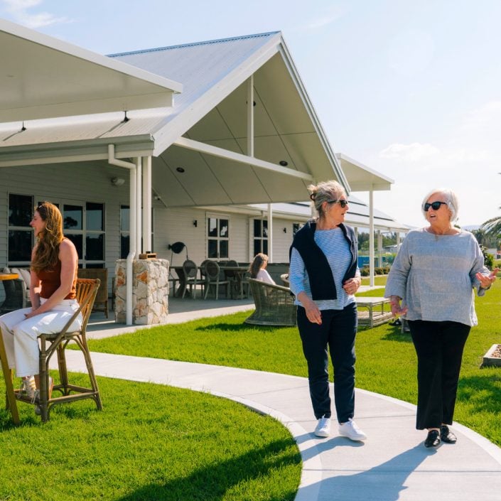 Two women stroll and chat along a garden path outside a modern house, while a man and woman enjoy conversation at a patio table—capturing the relaxed lifestyle of over-50s living in Hunter Valley under a sunny sky.