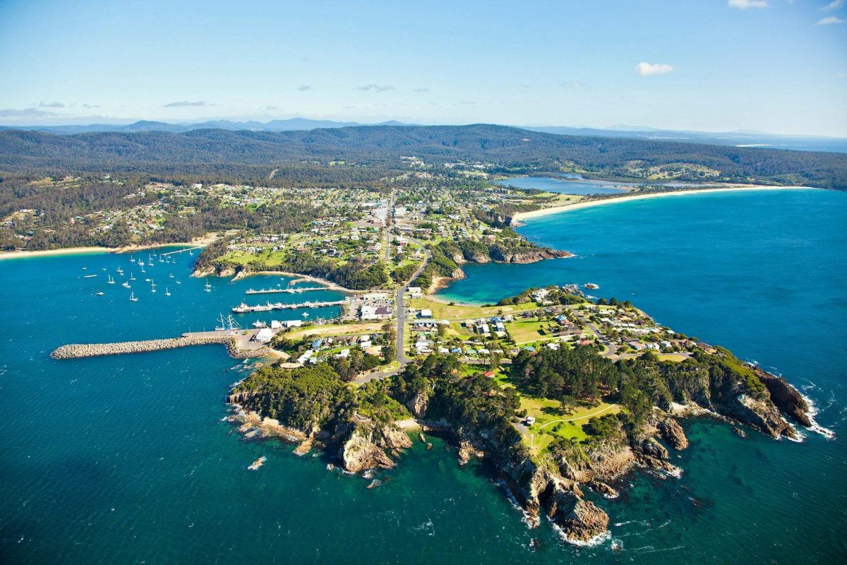 Aerial view of a coastal town on a peninsula surrounded by blue sea, with marina, green spaces, scattered houses including the Eden Gardens over 50s lifestyle community, sandy beaches, and forested hills under a clear sky.