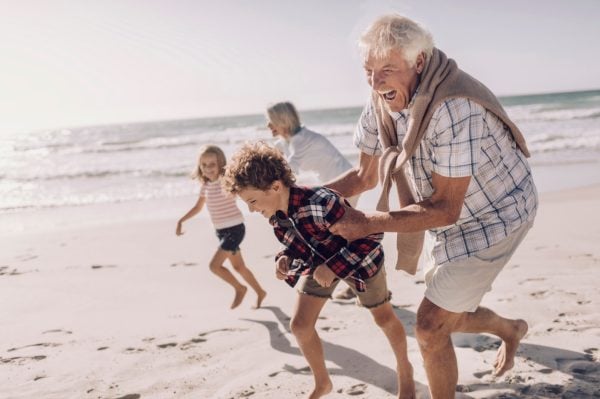 Two older adults and two children laugh and run along a sunny beach, embodying the joyful spirit of Eden Gardens over 50s lifestyle. The bright sand and sea backdrop highlight their playful moment together.