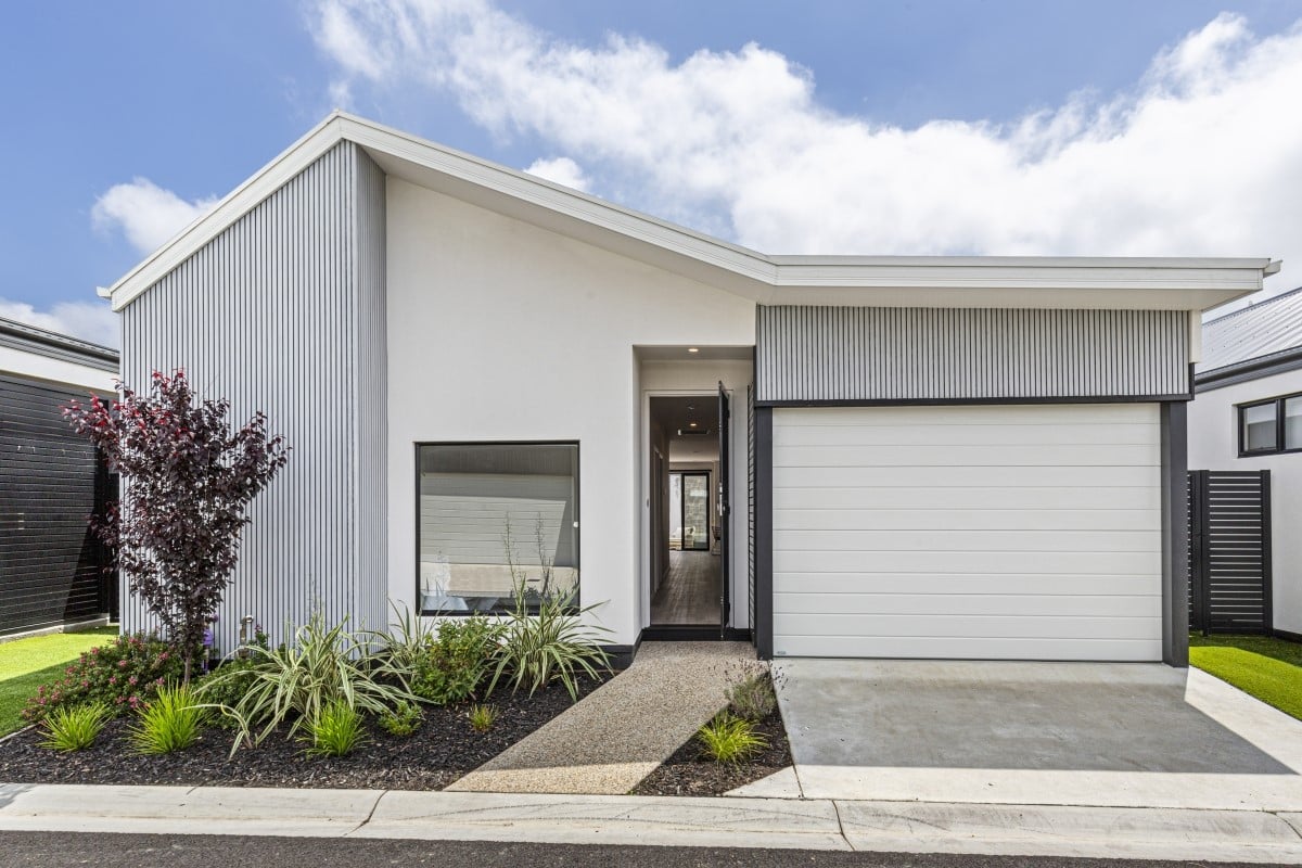 Modern single-storey house with a white exterior, grey accents, and a sloped roof. Featuring a garage, large window, and landscaped garden with shrubs and grass under a partly cloudy sky. Open for Viewing—come explore this inviting home!.
