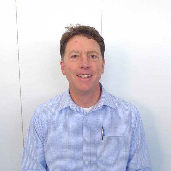 Chris Wilson, a middle-aged man with short brown hair, stands against a plain white background in a light blue chequered shirt, smiling at the camera. A pen peeks out from his shirt pocket.