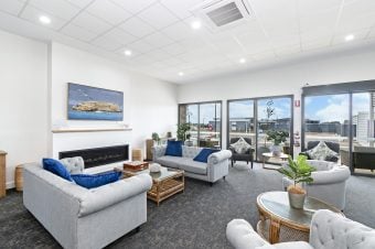 A modern lounge designed for land lease living, featuring grey sofas, blue and white cushions, a fireplace, large windows with natural light, indoor plants, and a framed seascape painting on the wall—perfect for relaxing retirement.