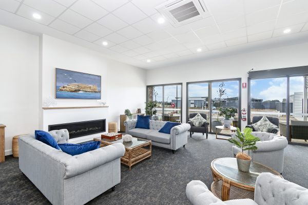 A modern lounge designed for land lease living, featuring grey sofas, blue and white cushions, a fireplace, large windows with natural light, indoor plants, and a framed seascape painting on the wall—perfect for relaxing retirement.