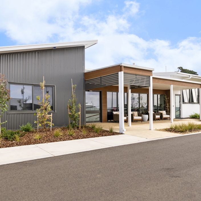 A modern, single-storey building with metal and white panelling, large windows, and a covered patio offers stylish land lease living for retirement amid landscaped plants and a paved drive under a partly cloudy sky.