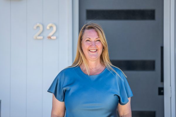 A smiling woman with long blonde hair stands in front of a modern grey door and a white wall with the number 22 displayed beside her. She is wearing a short-sleeved blue top.
