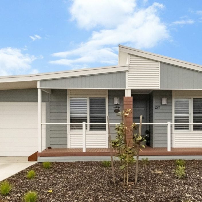 Single-storey modern house with grey and white exterior, a covered front porch, white railing, garage on the left, and landscaped garden—perfect for those seeking Edgewater accommodation—under a blue sky with clouds.