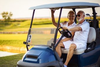 An older couple, both wearing white clothing and sunglasses, smile whilst sitting together in a blue golf buggy on a sunny golf course. The woman hugs the man as he holds the steering wheel.