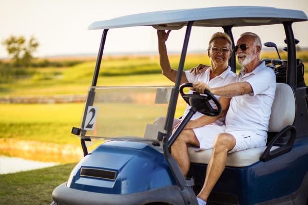 An older couple, both wearing white clothing and sunglasses, smile whilst sitting together in a blue golf buggy on a sunny golf course. The woman hugs the man as he holds the steering wheel.
