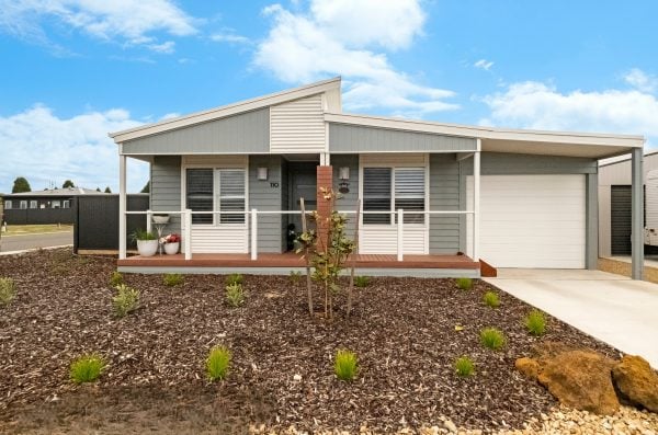 Single-storey modern house on Lot 15 in Edgerton with a covered front porch, garage, and drive. The garden features mulch, small green plants, and rocks under a blue sky with some clouds.