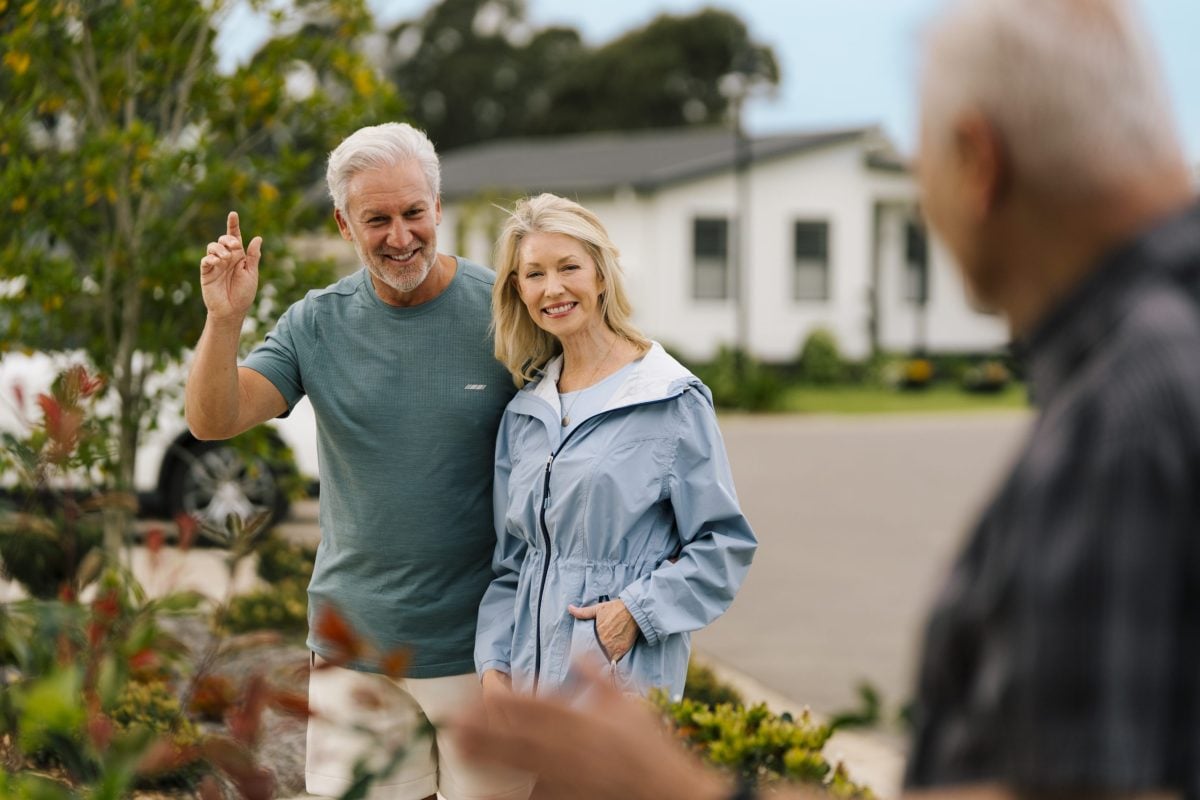 A smiling older couple walks outdoors in a residential neighbourhood, enjoying land lease living in retirement. They wave to another older man greeting them from the foreground, with trees and houses visible in the background.