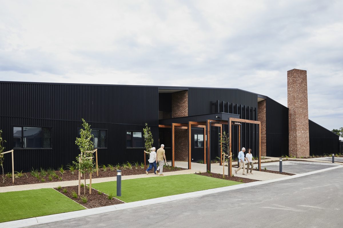 A modern black building with angular lines, brick accents, and a tall chimney offers inviting retirement living. Several people walk the path towards the entrance, framed by young trees along the landscaped grounds.