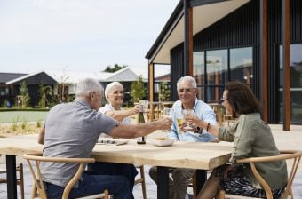 Four older adults enjoy retirement living as they sit around a wooden outdoor table, smiling and clinking glasses together over food and drinks. Modern houses and lush greenery provide a beautiful backdrop to their gathering.