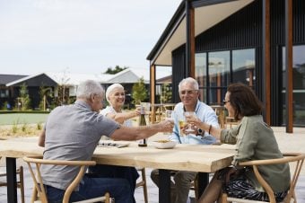 Four older adults enjoy retirement living as they sit around a wooden outdoor table, smiling and clinking glasses together over food and drinks. Modern houses and lush greenery provide a beautiful backdrop to their gathering.