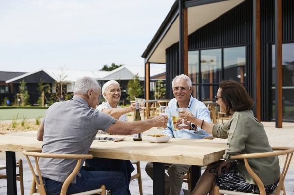 Four older adults enjoy retirement living as they sit around a wooden outdoor table, smiling and clinking glasses together over food and drinks. Modern houses and lush greenery provide a beautiful backdrop to their gathering.