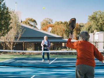 Two older adults enjoy a game of pickleball on an outdoor court at Lincoln Lifestyle Baranduda, an ideal spot for the over 50s. One in an orange jumper hits the ball, while the other stands ready. Trees and a building complete the scene.