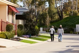 An elderly couple walks together down a quiet, sunny residential street in an over-50s community in Bendigo, with landscaped gardens, houses on one side, and trees in the background.