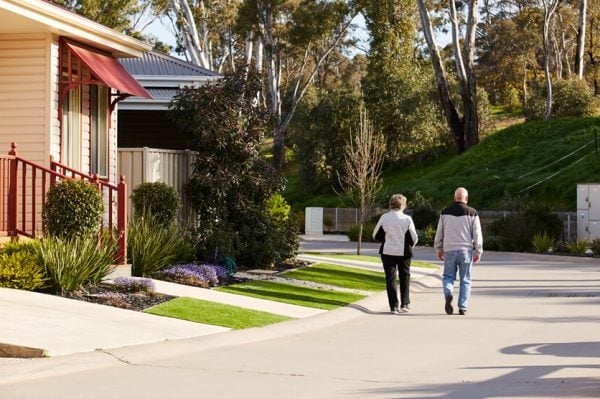 An elderly couple walks together down a quiet, sunny residential street in an over-50s community in Bendigo, with landscaped gardens, houses on one side, and trees in the background.