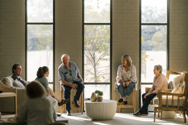 A group of five older adults from Lincoln Lifestyle Baranduda over 50s sit together in a bright, sunlit room with large windows, chatting and smiling. The space is furnished with comfortable chairs and a coffee table with potted plants.