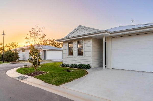 Single-storey modern house with light-coloured cladding, a white garage door, and a small landscaped front garden—perfect for over-50s living in Northern Rivers. Set on a paved street at sunset, with trees and another similar house in the background.