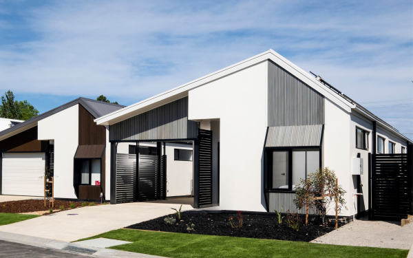 A modern, single-storey Seattle SX house with a sloping roof, white and grey exterior, large windows, and a neat front garden with green grass and minimal landscaping under a blue sky.