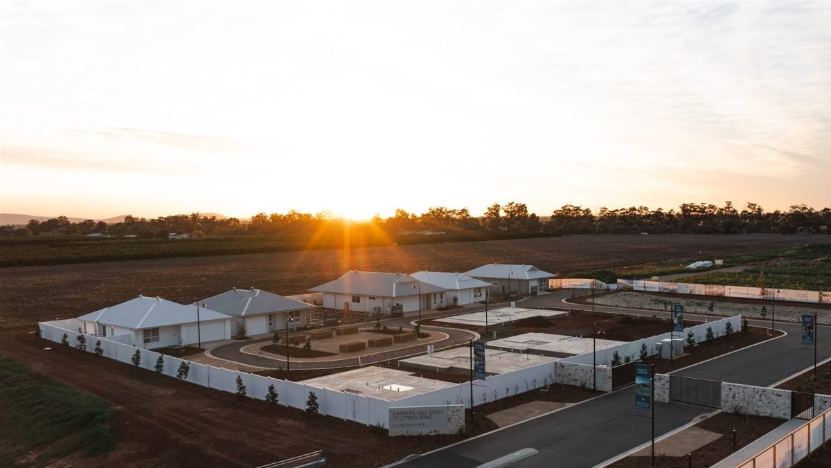 A small cluster of modern white houses sits within a fenced area, surrounded by open fields at sunset—an ideal setting for over-50s living in Griffith, where the glowing sky casts long shadows across a peaceful neighbourhood.