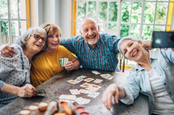 Four older adults smiling and taking a selfie together while playing cards at a table. As poker chips and cards are spread out in front of them, they enjoy a friendly downsizing conversation in the bright daylight streaming through large windows.