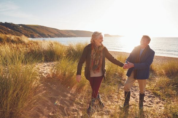 An older couple walk hand in hand through grassy dunes by the beach, smiling at each other as the sun sets over the sea in the background—come explore our communities and create your own cherished moments.