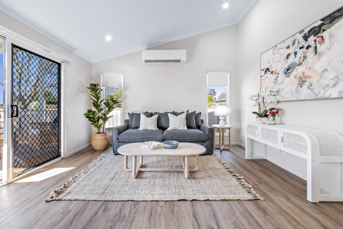 Modern living room at Sundown Lifestyle Estate with a grey sofa, neutral cushions, and a round coffee table on a woven rug. Large windows and glass door invite natural light; stylish decor features plants, mirrors, and abstract art on white walls.