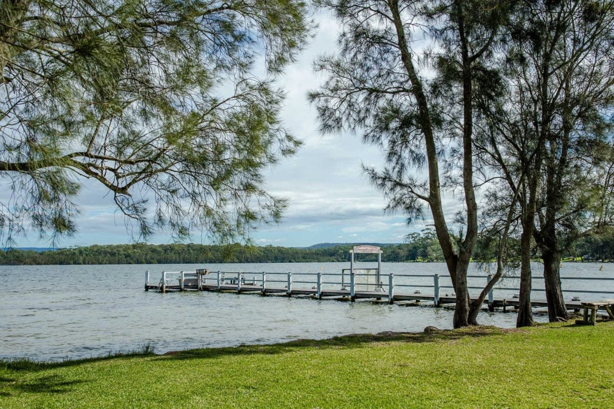 A wooden jetty extends over a calm lake at Rosevale Home Village, surrounded by green grass and tall trees. The sky is partly cloudy, with distant hills visible across the water.