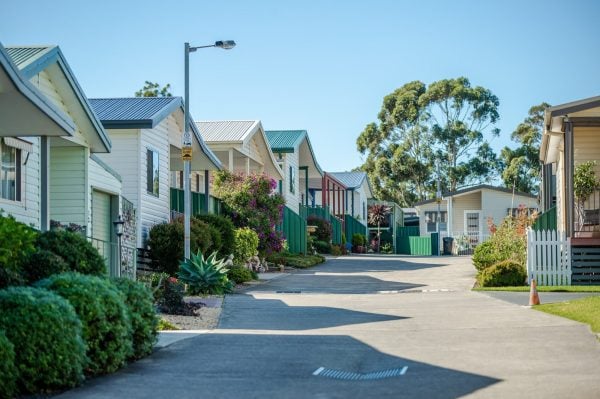 A sunny, quiet residential street in Rosevale Home Village is lined with small, colourful houses and neatly trimmed bushes, set against a clear blue sky and tall trees in the background.