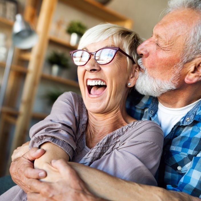 An older couple sit together, laughing joyfully. The woman, wearing glasses, smiles widely whilst the man in a checked shirt hugs her from behind and kisses her cheek. They appear happy and relaxed in a cosy home, perhaps after using a downsizing calculator.