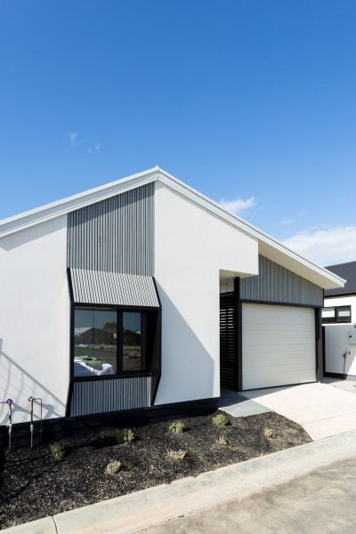 Modern single-storey house with a white exterior, black trim, and geometric design. There is a single-car garage, large window, small landscaped front garden, and a clear blue sky overhead.