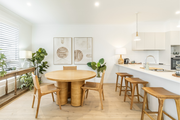 Bright, modern dining and kitchen area in Lot 220 with a round wooden table, four chairs, bar stools at a white counter, potted plants, abstract wall art, and light wood flooring—a perfect Bancroft retreat.