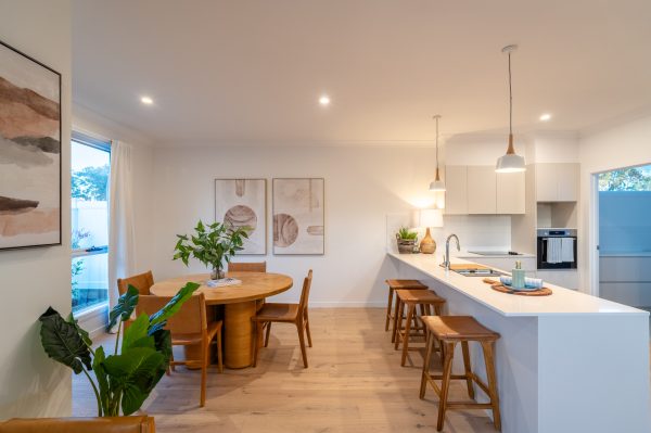 Modern kitchen and dining area in Lot 250, Bancroft, featuring light wood floors, a round dining table with chairs, white worktops, pendant lights, plants, and minimalist decor. Large windows flood the space with natural light.