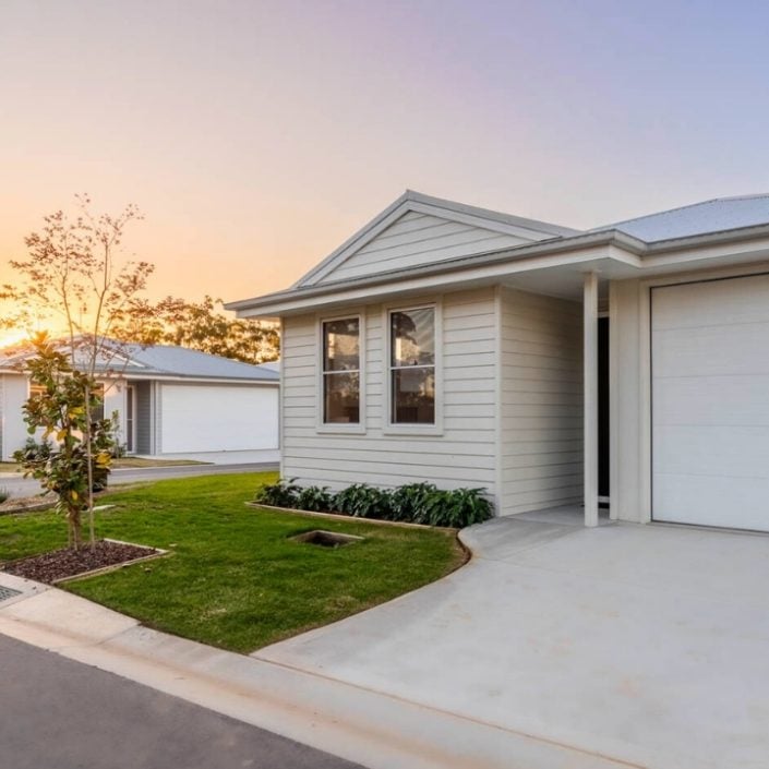 A modern single-storey house on Lot 250 with light-coloured siding, a garage, and a neatly landscaped front garden at sunset. Situated in the tranquil Bancroft neighbourhood, similar homes and a quiet street are visible in the background.
