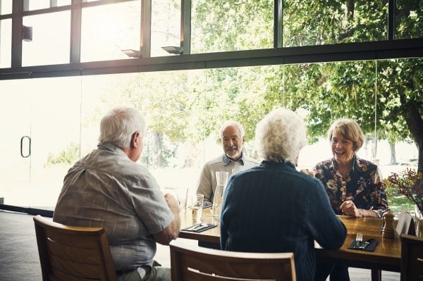 Four older adults sit together at a dining table in a bright, modern restaurant with large windows, enjoying a meal and conversation after an engaging Information Session, with trees visible outside in the sunlight.