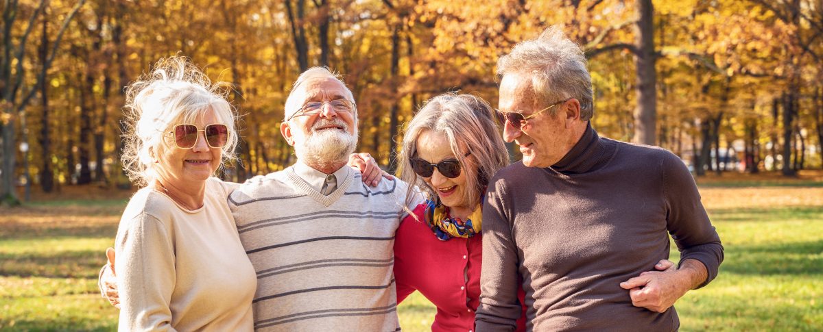 Four older adults wearing sunglasses stand close together, smiling and enjoying a sunny day in a park with autumn trees, celebrating the official launch of autumn with golden leaves in the background.