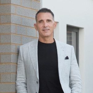 Michael Musitano, in a light grey blazer and black shirt, stands casually against a brick corner outside, smiling slightly at the camera.