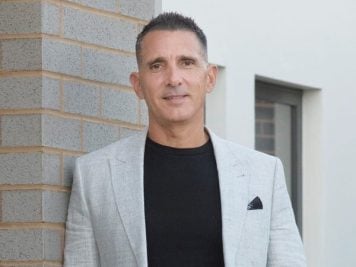 Michael Musitano, in a light grey blazer and black shirt, stands casually against a brick corner outside, smiling slightly at the camera.