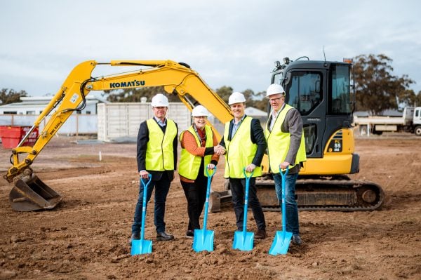 Four people in helmets and yellow safety vests stand on a construction site for Huntly Lifestyle Estate, each holding a blue spade at the Sod Turning ceremony. A yellow Komatsu excavator is behind them on the dirt ground.