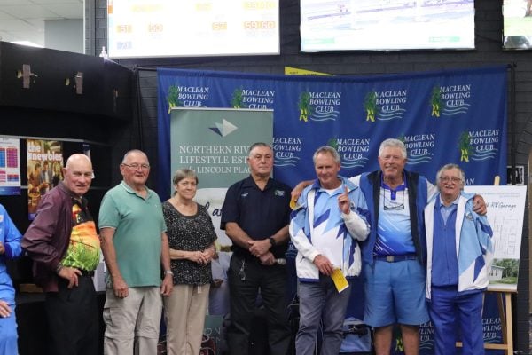 A group of eight adults stands indoors, some in blue sports kits, posing in front of a “Maclean Bowling Club” banner at Northern Rivers Lifestyle Estate. They are smiling, and one man is holding up two fingers in a peace sign.
