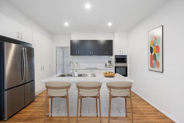 Modern Emerson kitchen with a large island, three beige barstools, stainless steel fridge, oven, and microwave, white cupboards, wooden floor, and abstract art on the wall—stylish living at Lot 41.